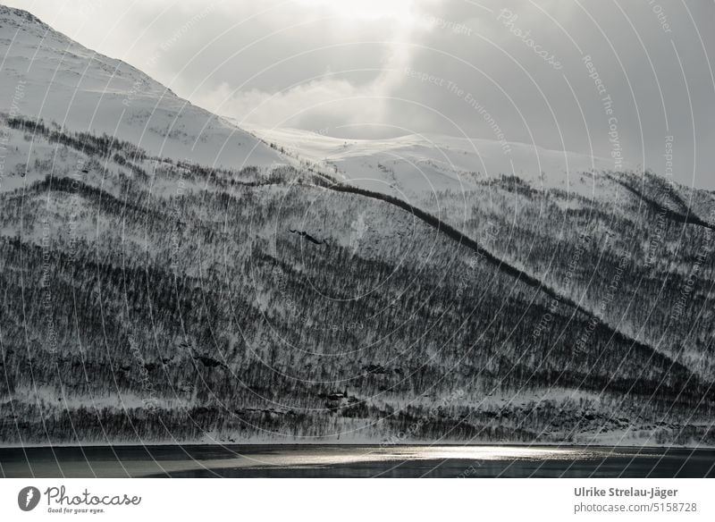 Einsames Haus am Fjord in Abendstimmung mit schneebedecktem Berghang, Wald und Sonne hinter Wolken einsames Haus Winter Wintertag Schnee kalt Winterstimmung