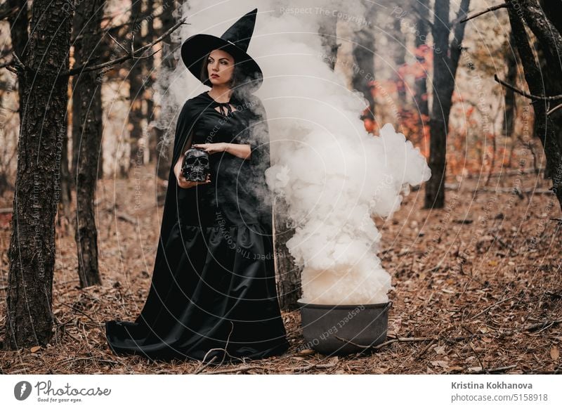 Schwarze Hexe hält einen Totenkopf in den Händen und streichelt ihn auf einem herbstlichen Waldhintergrund. Alchemie wütend Kunst Herbst Hintergrund schlecht