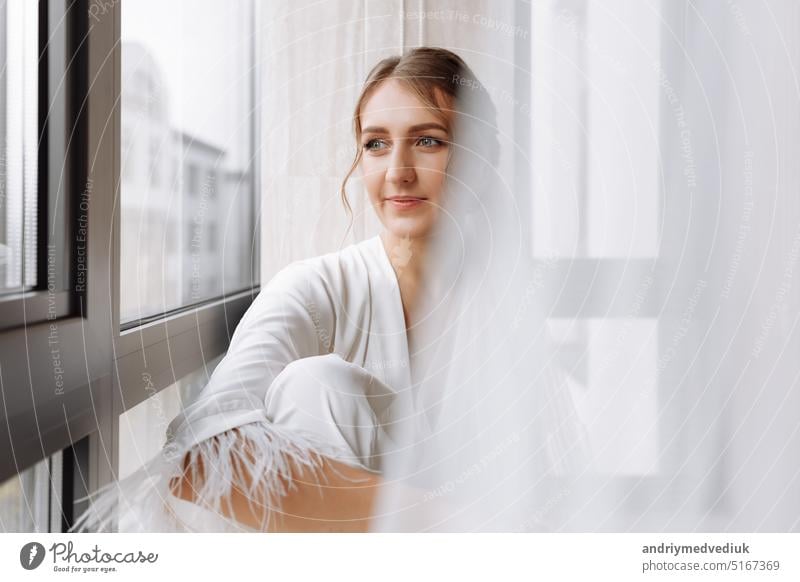 Braut s Morgen. Braut trinken Champagner in der peignoir. junge Frau sitzt auf einem großen Fenster in einem Hotelzimmer im Bad. Schönes Mädchen in weißen Hochzeitsgewand. Hochzeitstag