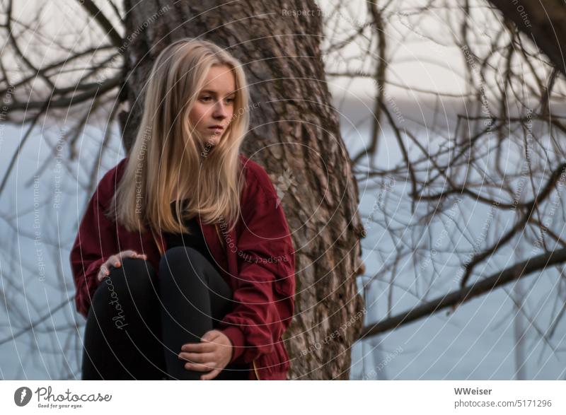 Ein junges blondes Mädchen sitzt nachdenklich auf einem schief wachsenden Baum am Seeufer junge Frau Natur draußen introvertiert sensibel schön traurig ratlos