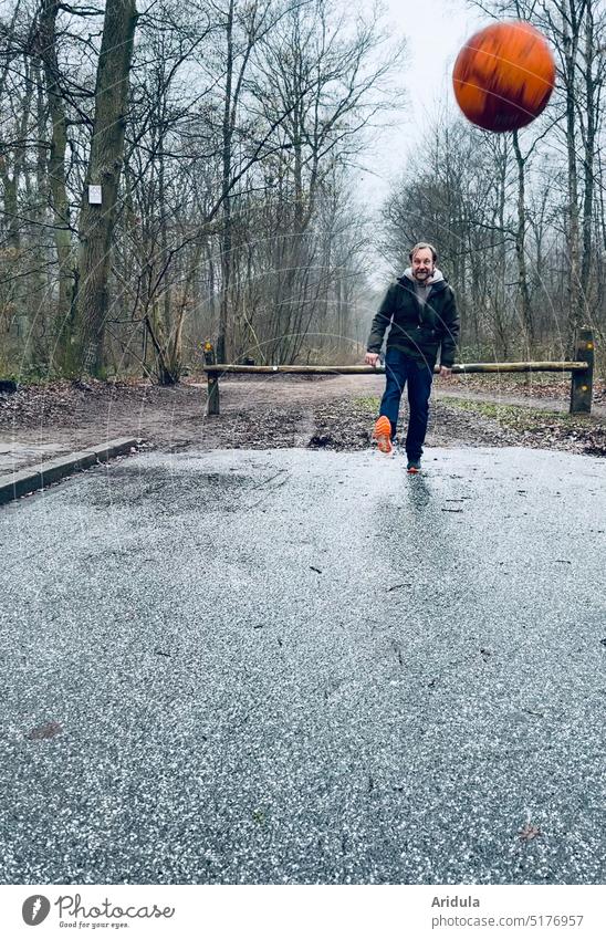 Mann steht am Waldrand auf Asphalt und schießt orangenen Fußball in Richtung Kamera schießen Bewegung Sport Ball Freude Außenaufnahme Freizeit Winter Bäume