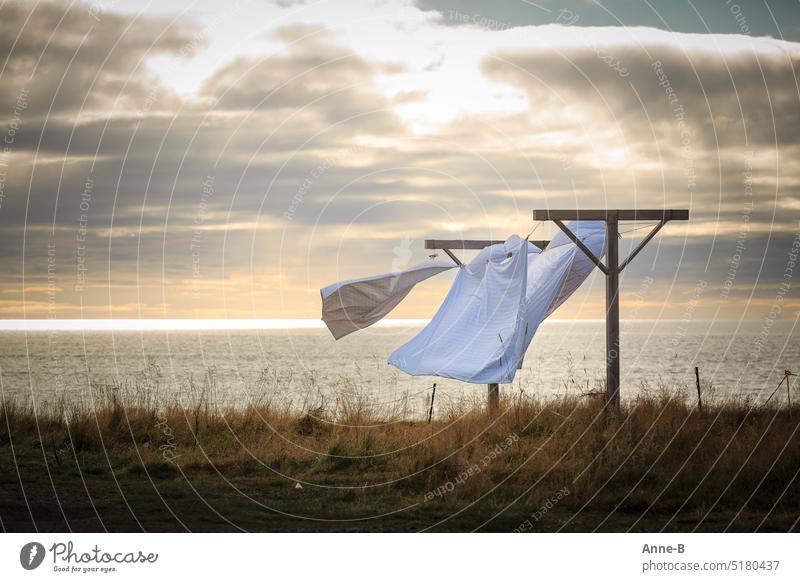 Weiße Bettwäsche wedelt im Wind auf einer Wiese direkt am Meer bei wunderbarem Gegenlicht und leicht bewölktem Himmel. Wäsche Leine draußen trocknet Wetter