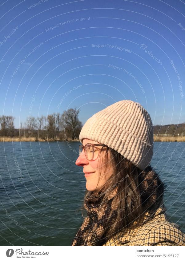 eine frau mit mütze und brille steht mit geschlossenen augen an einem  fluss und genießt die sonnenstrahlen inn meditation geschlossene augen genießen wohltuend