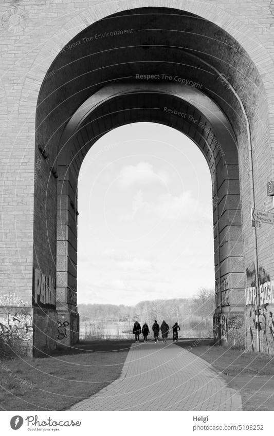 unter dem Bogen eines Viadukts sind 5 nicht erkennbare, scheinbar winzige Menschen  unterwegs, 4 zu Fuß, einer per Fahrrad Bauwerk Architektur Silhouette