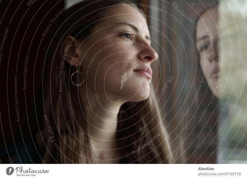 Portrait einer jungen, brünetten Frau mit schulterfreiem Pullover, die vor einer roten Wand am Fenster steht und deren Antlitz sich in der Scheibe spiegelt