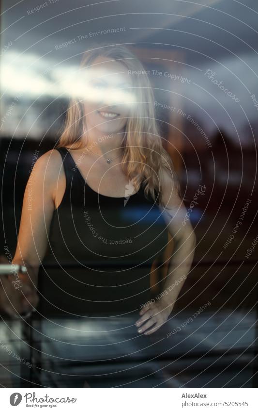 Portrait einer blonden, langhaarigen Frau in schwarzem Tanktop hinter einer Fensterscheibe lächeln Blick in die Kamera schlank anmutig schön natürlich