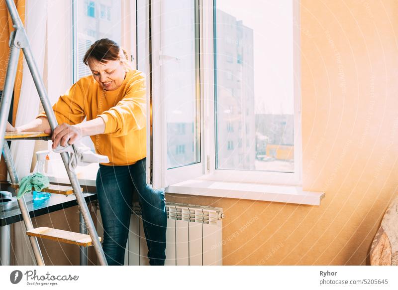 Lächelnde kaukasische Frau von fünfzig in gelbem Pullover und Jeans wäscht staubige Fenster in der Wohnung. 50 Jahre alte Frau reinigt Fenster von Flecken mit Lappen und Sprayreiniger. Ältere Frau reinigt Haus, macht Hausarbeit. Glückliche Frau, zufrieden mit...