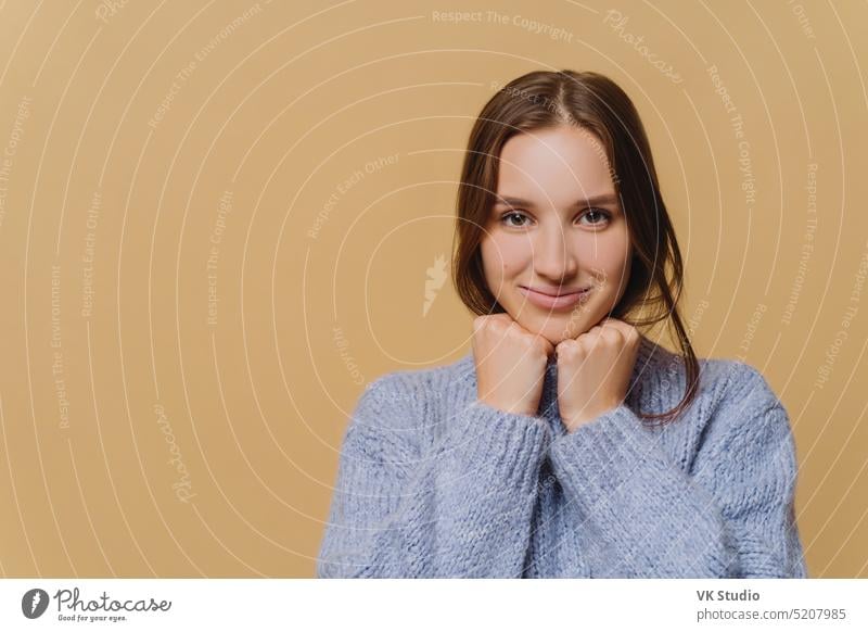 Junge europäische Frau mit minimalem Make-up, dunklen Haaren, bekleidet mit Winterpulli, hält die Hände unter dem Kinn, hat ein charmantes Lächeln, steht vor braunem Hintergrund, Textfläche für Ihren Werbeinhalt