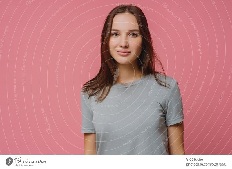 Adorable Millennial Mädchen mit glatten dunklen Haaren, trägt lässig t Shirt, schaut ernst in die Kamera, hat gesunde Haut, posiert über rosa Hintergrund. Angenehm aussehende Frau hat Gespräch mit jemandem.