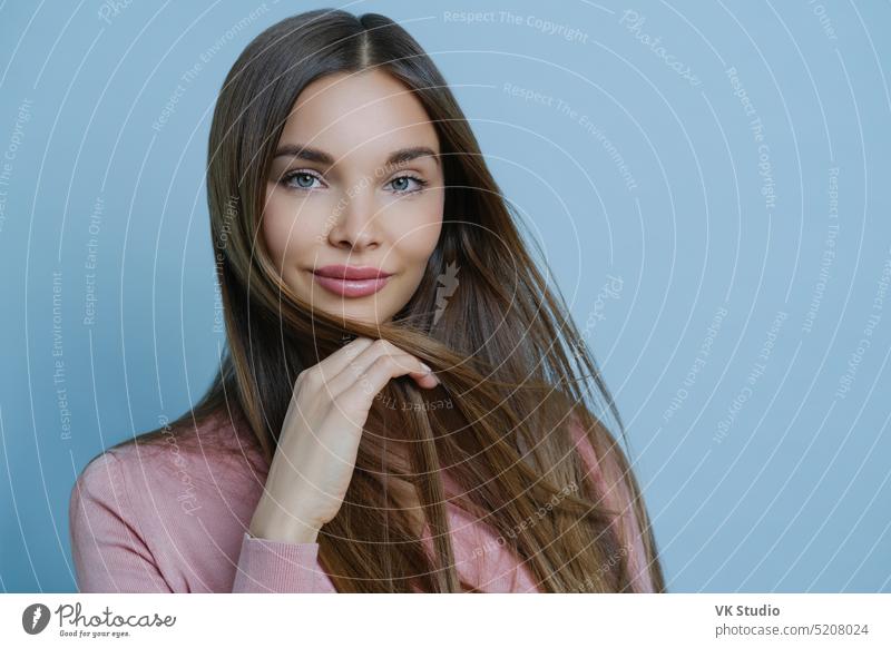 Close up shot von attraktiven weiblichen Modell zeigt ihre Sinnlichkeit und feminine Züge, hat gesunde leuchtende Haut, lange dunkle Haare, sieht mit ruhigen Gesichtsausdruck, isoliert auf blaue Wand. Schönheit und Gesundheit