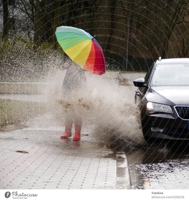 Wusch!!! Da donnert das dunkle Auto voll durch eine tiefe Pfütze. Ein riesiger Wasserschwall durchnässt die Fußgängerin mit einem bunten Regenschirm und roten Gummistiefeln.