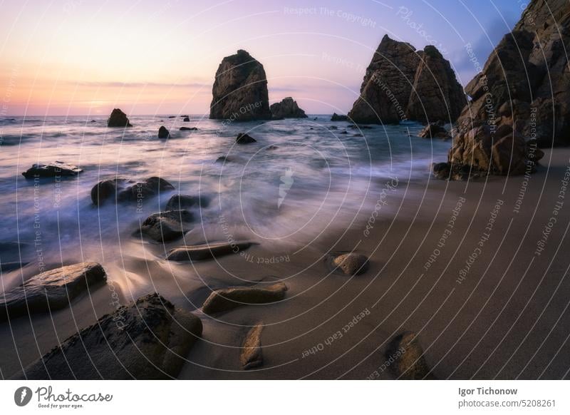 Malerische Küste von Ursa Beach, Sintra, Portugal. Sandstrand mit Felsklippen beleuchtet von Sonnenuntergang Abend weiches goldenes Licht. Atlantic Ocean Wellen in Langzeitbelichtung