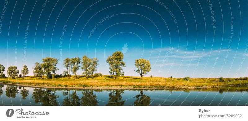 Panorama der blauen Nacht Sternenhimmel über See Fluss. Nacht glühenden Sternen und Bäumen Wälder im Wasser reflektiert. Schöne Nacht Landschaft mit natürlichen glühenden Sternen