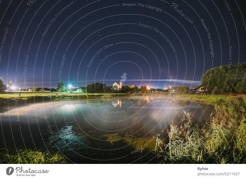 Nacht Wolken über Fluss See in der Nähe von Häusern im Dorf. Night Starry Sky Above Lake River With Bright Stars And Meteoric Track Trail. Glühende Sterne über Sommer Natur