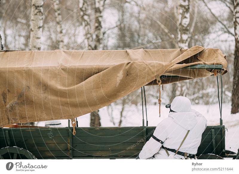 Hidden Re-enactor Dressed As German Wehrmacht Infanterie Soldat im Zweiten Weltkrieg Soldaten sitzen im Hinterhalt in der Nähe von Bauern Karren im Winter Wald und Blick auf alte Armee Ferngläser. Historisches Re-enactment. ZWEITER WELTKRIEG. Winter verschneiten Tag