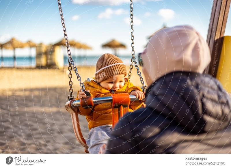 Mutter schiebt ihren Säugling auf einer Schaukel auf Sandstrand Spielplatz im Freien auf schönen sonnigen kalten Wintertag in Malaga, Spanien. Kind Glück Baby