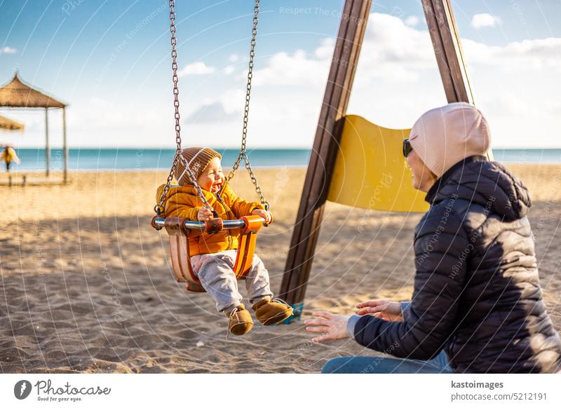 Mutter schiebt ihren Säugling auf einer Schaukel auf Sandstrand Spielplatz im Freien auf schönen sonnigen kalten Wintertag in Malaga, Spanien. Kind Glück Baby