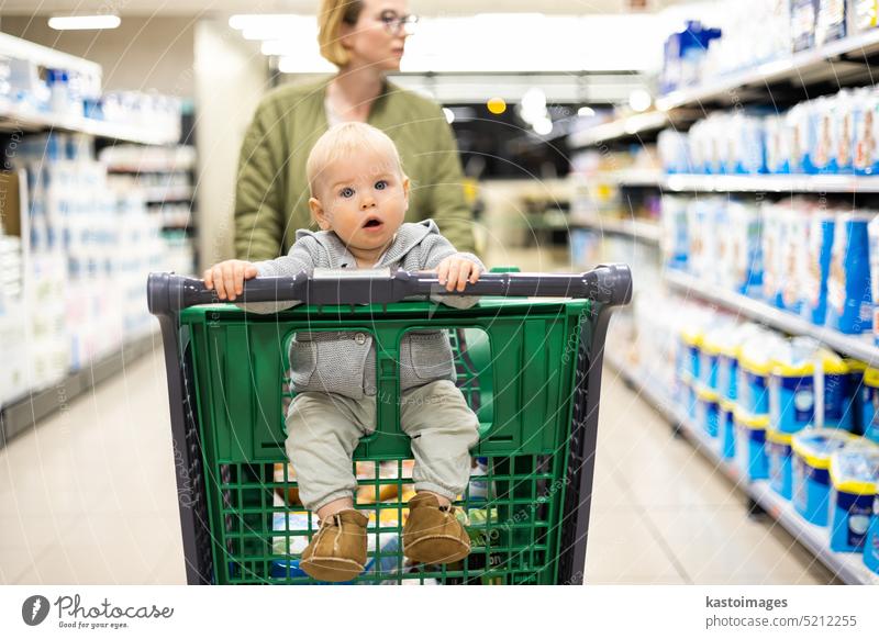 Mutter schiebt Einkaufswagen mit ihrem Säugling Baby Junge Kind nach unten Abteilung Gang im Supermarkt Lebensmittelladen. Einkaufen mit Kindern Konzept. Frau