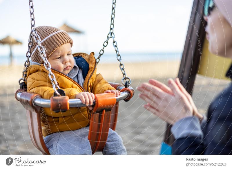 Mutter schiebt ihren Säugling auf einer Schaukel auf Sandstrand Spielplatz im Freien auf schönen sonnigen kalten Wintertag in Malaga, Spanien. Kind Glück Baby