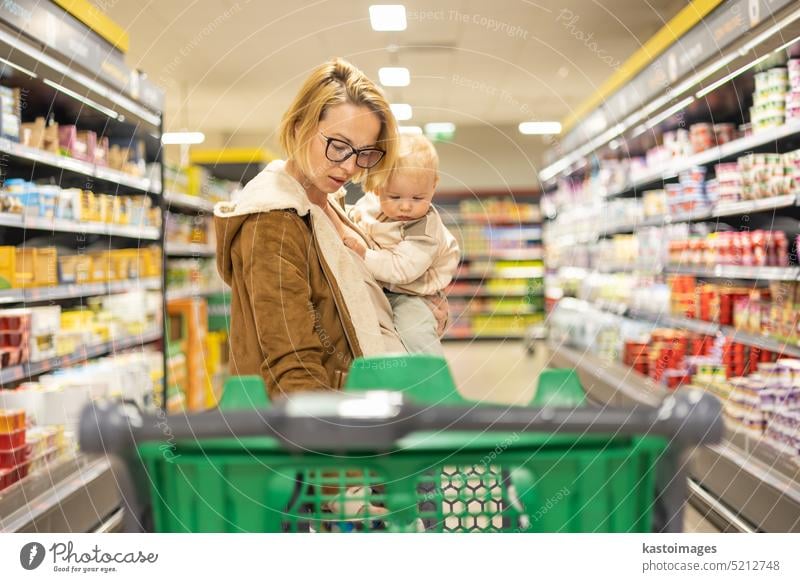 Eine Mutter kauft mit ihrem kleinen Jungen ein und hält das Kind, während sie an der Kasse im Supermarkt Produkte stapelt. Frau Baby Säugling Mama Laden kaufen