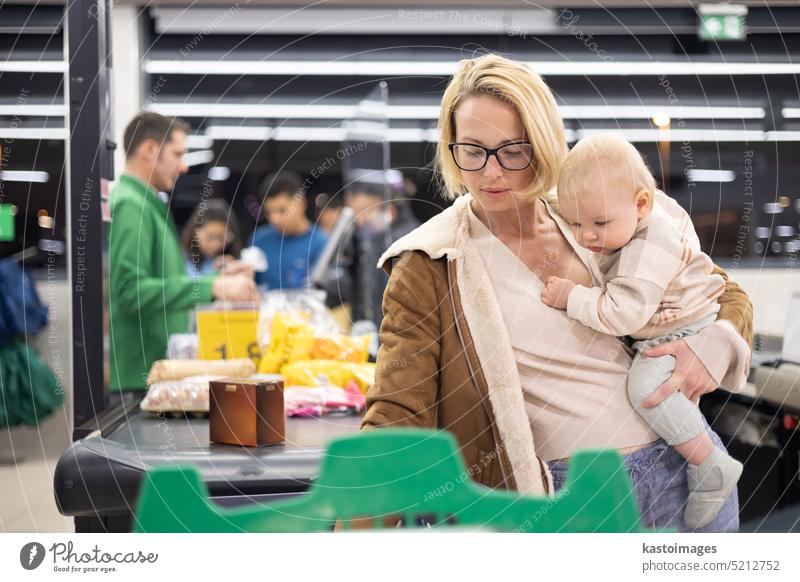 Eine Mutter kauft mit ihrem kleinen Jungen ein und hält das Kind, während sie an der Kasse im Supermarkt Produkte stapelt. Frau Baby Säugling Mama Laden kaufen