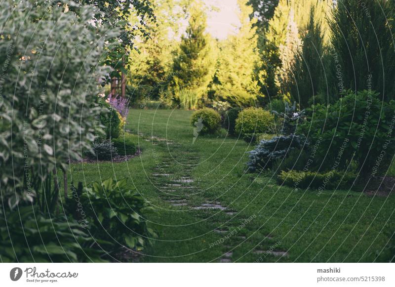 Abendlicher Sommerspaziergang im Garten mit geschwungenem Steinpfad und hölzernem Torbogen. Naturbelassener Waldgarten mit Hostas, Koniferen und Sträuchern