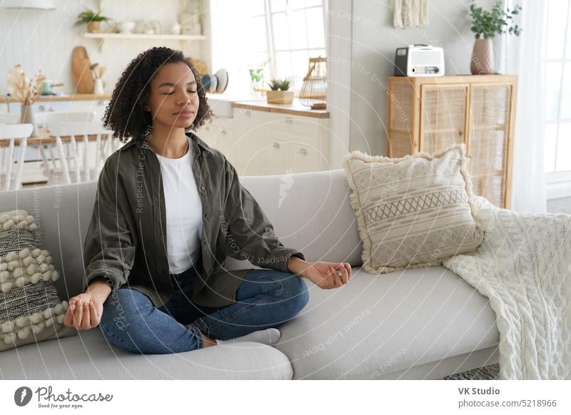 Ruhig gemischte Rasse Mädchen macht Yoga zu Hause, sitzen in Lotus-Pose auf dem Sofa. Meditation, gesunder Lebensstil meditieren heimwärts Wellness Windstille