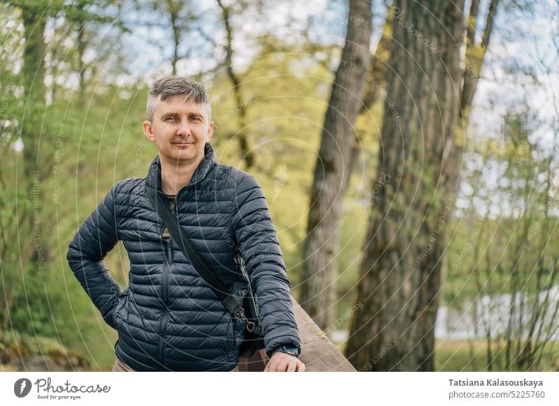 Ein Mann mittleren Alters mit grauem Haar in einer dünnen Daunenjacke im Vorfrühling, umgeben von Bäumen Wald Park Frühling Natur Porträt Lifestyle im Freien