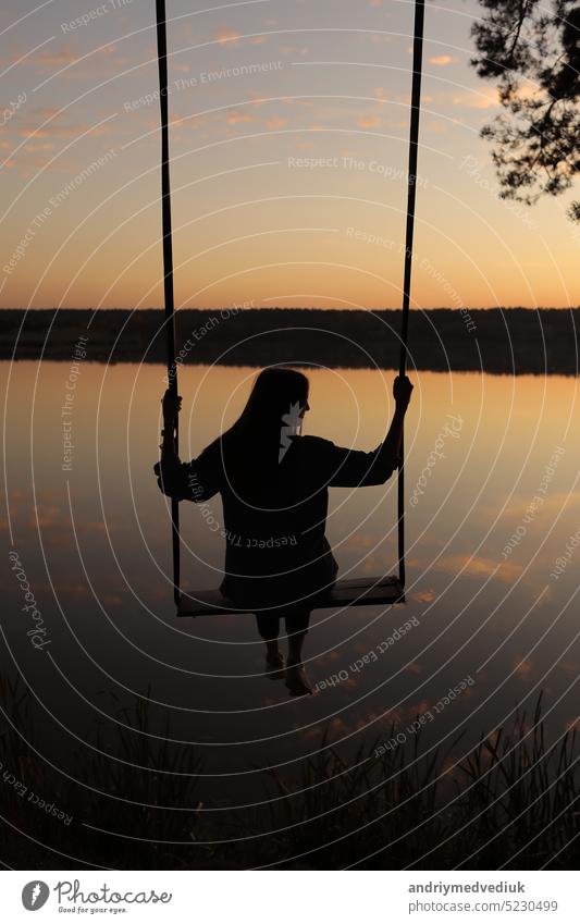 Silhouette einer romantischen jungen Frau auf einer Schaukel über einem See bei Sonnenuntergang. Junges Mädchen Reisende sitzen auf der Schaukel in der schönen Natur, Blick auf den See