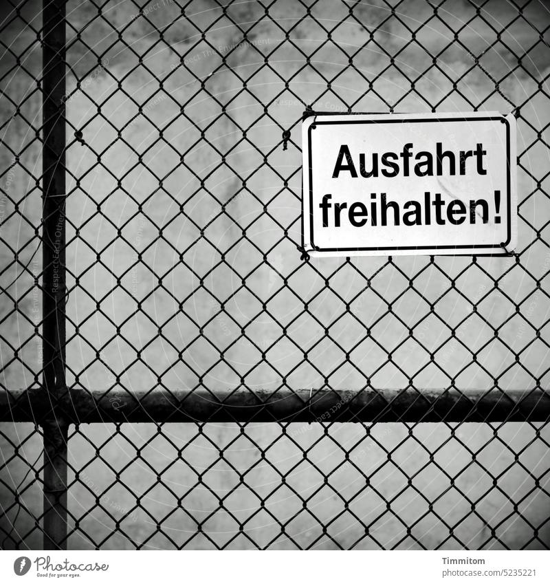 Ausfahrt freihalten! Schilder & Markierungen Hinweisschild Schriftzeichen Pfosten Metall Zaun Maschendrahtzaun trist grau Schwarzweißfoto Menschenleer