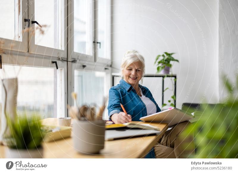 Lächelnde reife Geschäftsfrau, die in ein Notizbuch schreibt, während sie am Tisch im Büro sitzt echte Menschen Senior im Innenbereich Loft Fenster heimwärts