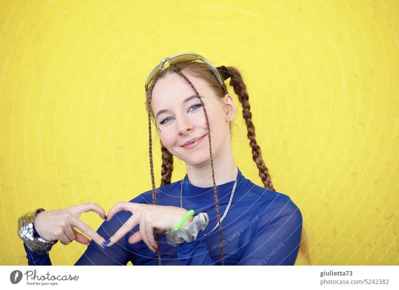 Stylisches Fashion Girl mit cooler Frisur und blauem Shirt, mit den Fingern ein Herz zeigend vor gelbem Hintergrund Porträt Lächeln positiv Frieden Liebe Glück