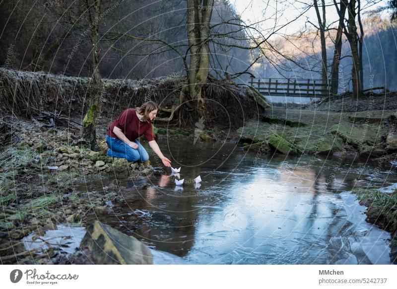 Frau sitzt am Ufer und lässt Papierschiffchen ins Wasser Ziel minimalistisch feminin Lächeln Teich See unanhängig dunkel Sympathie positiv Glück Freiheit