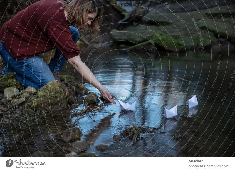 Frau sitzt am Ufer und lässt Papierschiffchen ins Wasser Ziel unanhängig See Teich dunkel Glück positiv Sympathie Freiheit minimalistisch feminin Lächeln