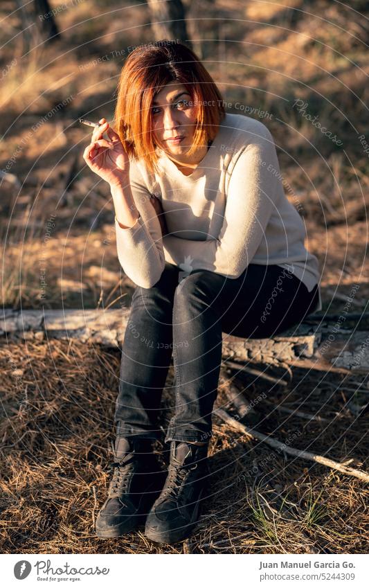 Junge Frau mit intensivem und zuversichtlichem Blick, rauchend auf einem Waldstamm in der Abenddämmerung sitzend Ermächtigung Selbstvertrauen Natur Stärke