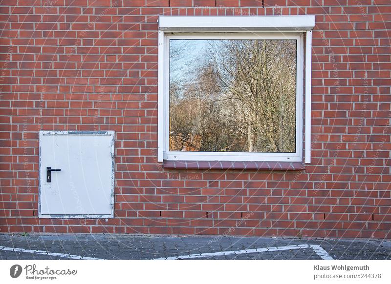 Eine verklinkerte Wand mit großem Fenster in dem sich Bäume und der Himmel spiegeln.Eine kleine Tür macht neugierig. Womöglich leben hier Liliputaner? Fassade