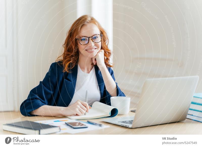 Erfolgreicher Wirtschaftswissenschaftler in Brille, prüft Informationen auf Laptop, liest Informationen auf Finanz-Website, schreibt in Notizblock und trinken heißen Kaffee, sitzt am Arbeitsplatz. Geschäft und Arbeit Konzept