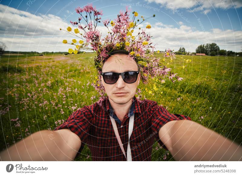 Junger lächelnder Mann mit Wildblumenkranz und Sonnenbrille macht ein Selfie mit einer Weitwinkelkamera und hat Spaß auf dem Feld an einem Sommertag. Positive Kerl hat keine Allergie. Lifestyle Sommer Konzept.