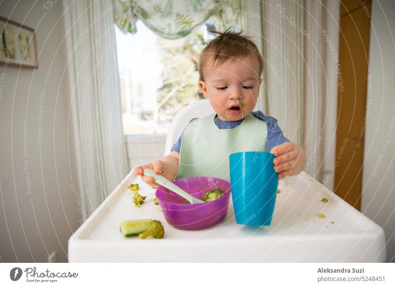 Süßes Baby isst erste feste Nahrung, Kleinkind sitzt im Hochstuhl. Kind probiert Gemüse am Tisch, entdeckt neue Lebensmittel. Gemütliche Küche Interieur. Gesundes Essen Konzept.