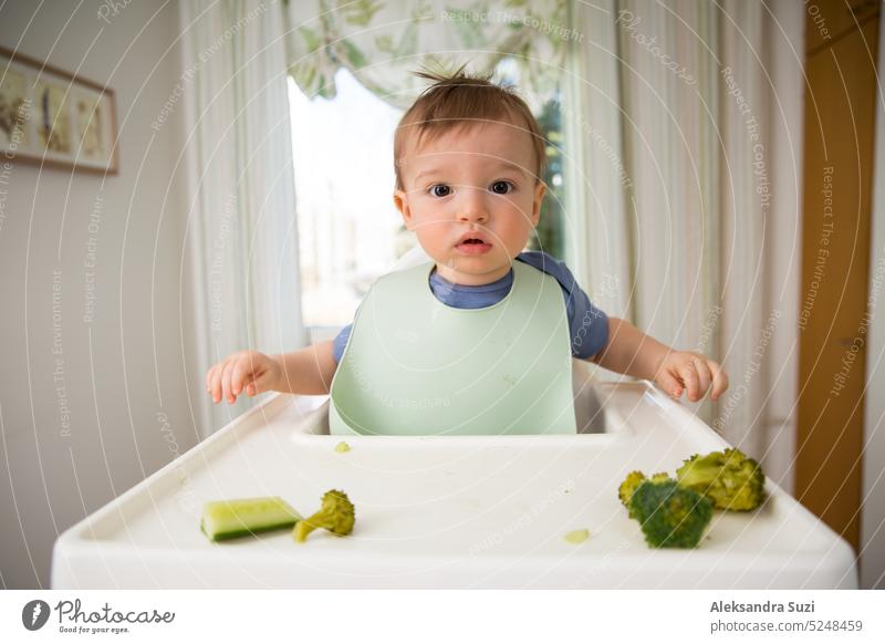 Süßes Baby isst erste feste Nahrung, Kleinkind sitzt im Hochstuhl. Kind probiert Gemüse am Tisch, entdeckt neue Lebensmittel. Gemütliche Küche Interieur. Gesundes Essen Konzept.