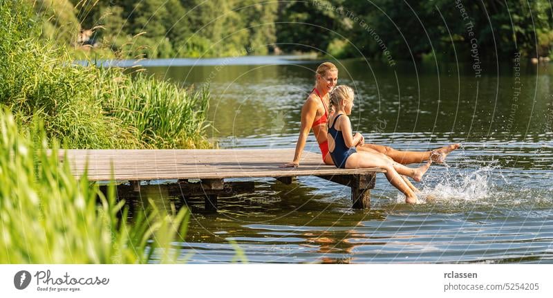 Mutter und Tochter baden und spritzen mit dem Fuß am Steg eines Sees im Sommer ins Wasser platschen Kind Schilfrohr Urlaub Badebekleidung Bikini Bayern