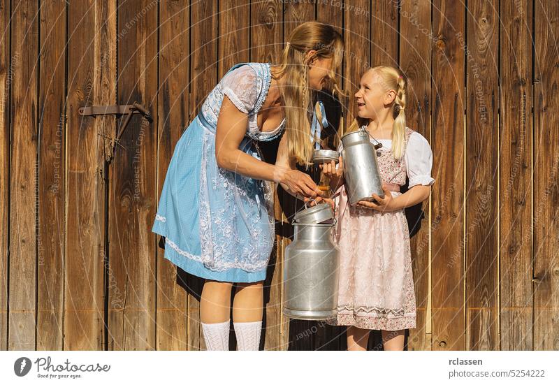 Mutter und Tochter halten Milchkannen in traditioneller bayerischer Tracht auf einem Bauernhof in Bayern auf einem Holzhintergrund. Oktoberfest Konzeptbild