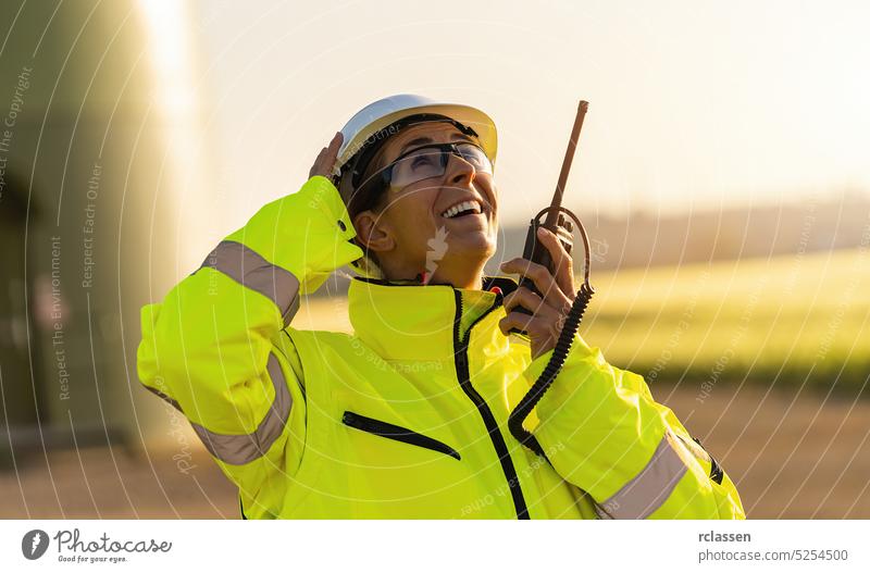 Ingenieurin mit Sicherheitsjacke steht vor einer Windkraftanlage und spricht in ein Walkie-Talkie, um die Windkraftanlage zu überprüfen Sicherheitsweste