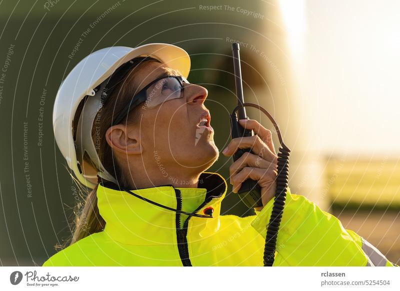 Eine Ingenieurin spricht skeptisch in ein Walkie-Talkie, um eine Windkraftanlage in einem Windpark zu überprüfen. Sicherheitsweste Maschinenbau Feld