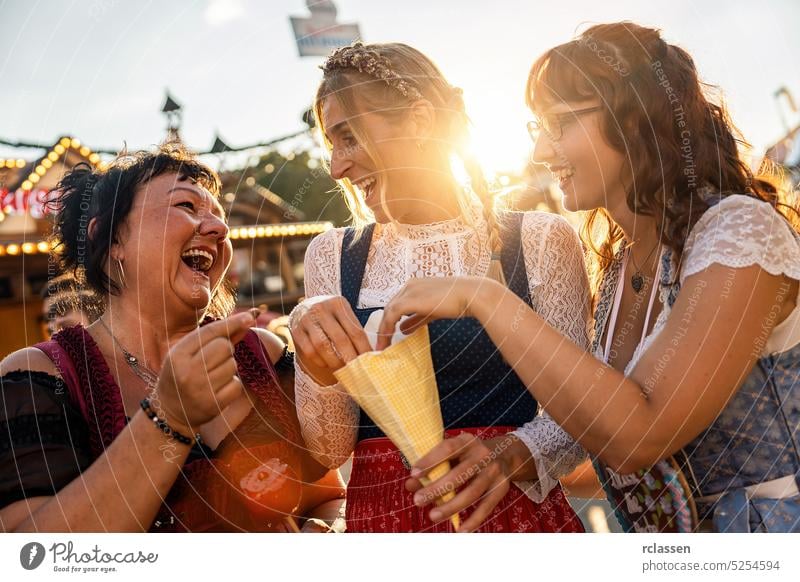 Fröhliche Freundinnen essen gemeinsam gebrannte Mandeln aus der Papiertüte vor dem Lebkuchenstand auf einer bayerischen Kirmes oder einem Oktoberfest oder dulden sich in Tracht oder Dirndl