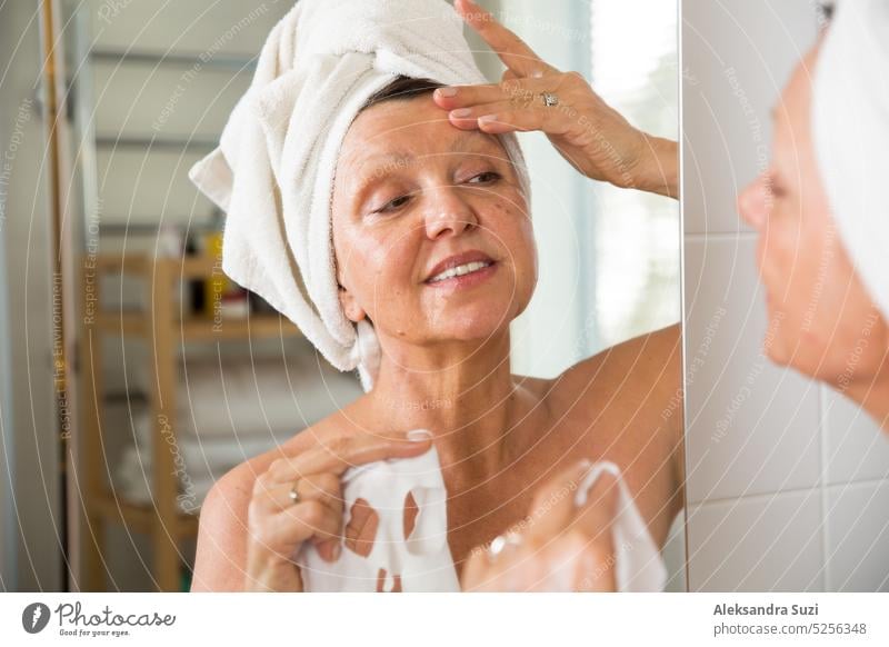 Reife schöne Frau Anwendung grün hausgemachte DIY Gesichtsmaske auf der Haut. Blick in den Spiegel im Bad, Spaß haben. Eingewickelt in ein Handtuch. Hautpflege-Routine und Anti-Aging-Behandlung für ältere Frauen.