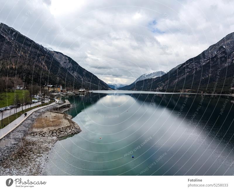 Blick über den Achensee in Österreich. In der Ferne liegt das Örtchen Jembach. Der See liegt ruhig da, umgeben von hohen Bergen. Der Himmel ist bewölkt.