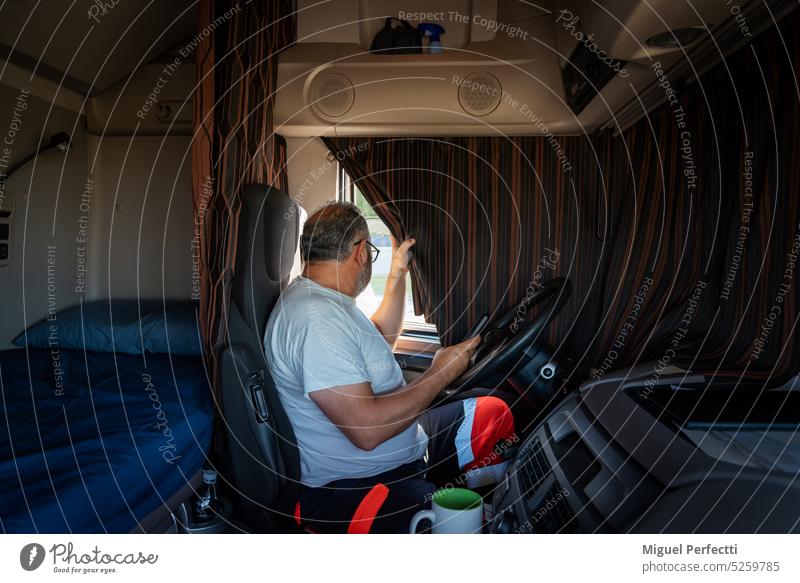 Ein älterer Lkw-Fahrer sitzt im Führerhaus seines Lkw und beobachtet bei zugezogenen Vorhängen, was draußen vor sich geht, während er auf eine Ladung wartet.