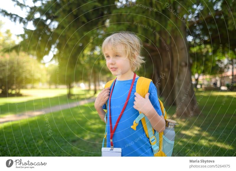 Grundschüler Junge mit Rucksack, Flasche Wasser und Namensschild an seinem Hals geht zur Schule. Glückliches Kind wartet auf den Schulbus. Schüler Tag elementar