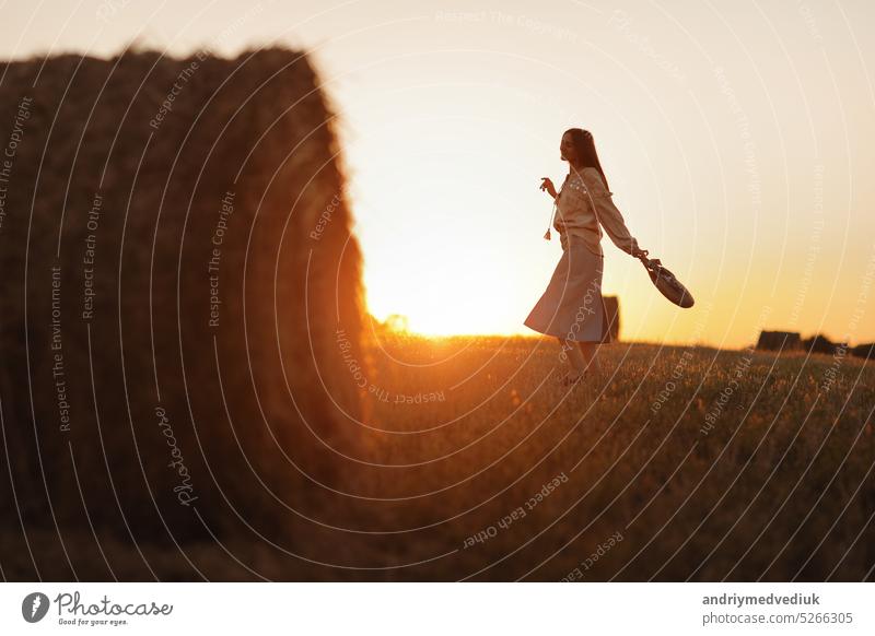 junge frau im schönen licht des sommerlichen sonnenuntergangs auf einem feld geht in der nähe der strohballen. schönes romantisches mädchen mit langen haaren im freien auf dem feld.